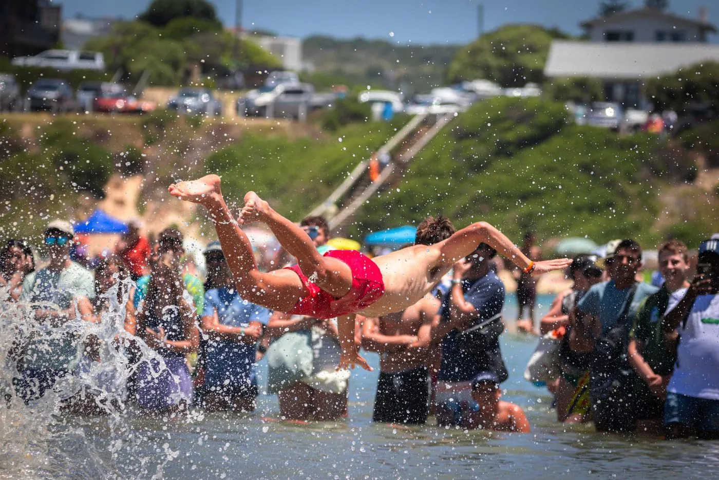 Players throw themselves into shallow water in front of a crowd on a beach.