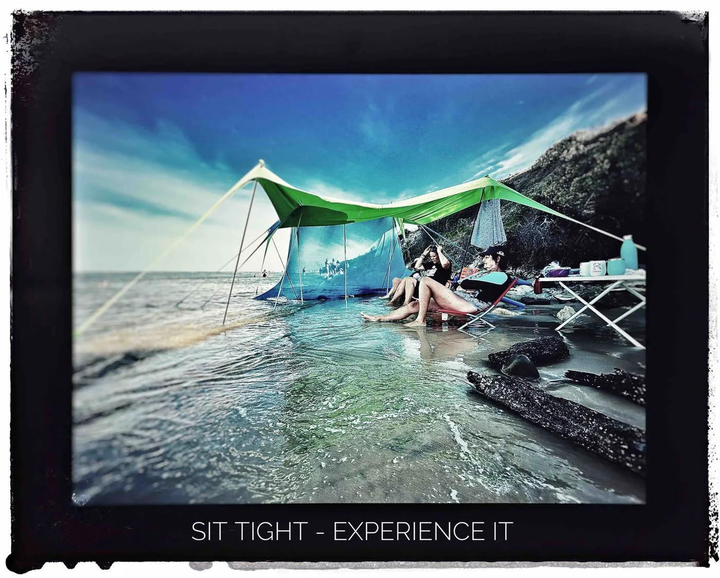 A green and blue shelter pitched over shallow water with a person seated beneath it, captioned Sit tight, experience it.