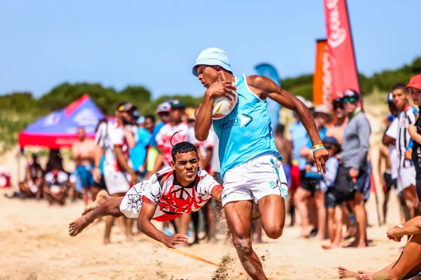 A rugby player carrying a ball is tackled on a sandy beach.