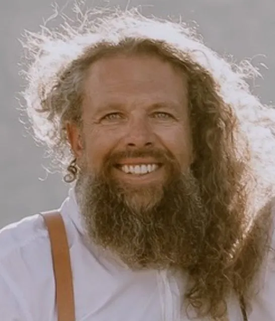 A smiling man with long wavy hair and a full beard, wearing braces.