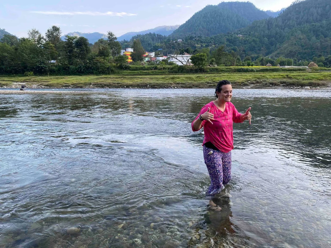 A woman in a pink top and dark trousers standing in a shallow river, with mountains and houses behind.