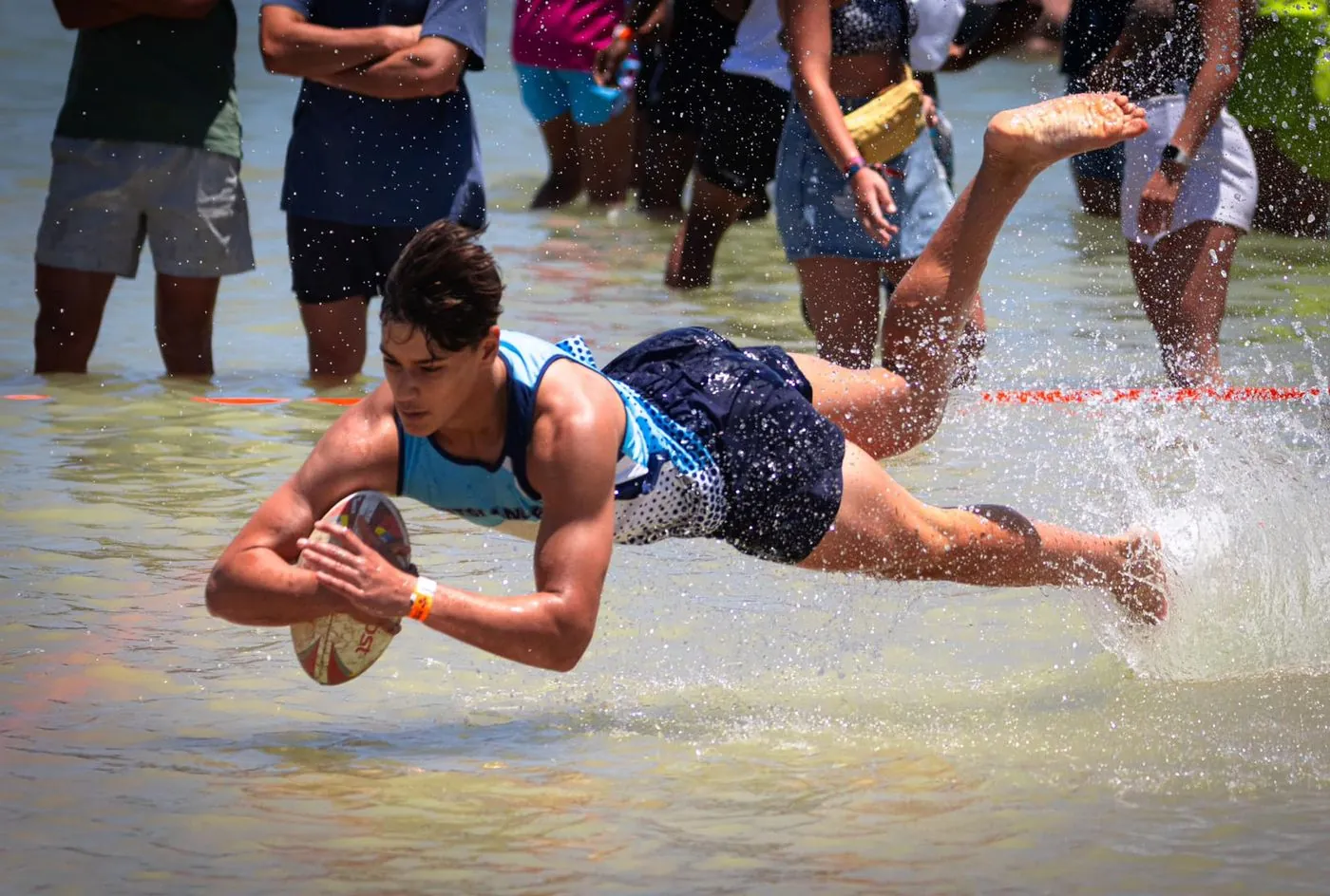 A player dives with a ball across shallow water while a crowd watches.