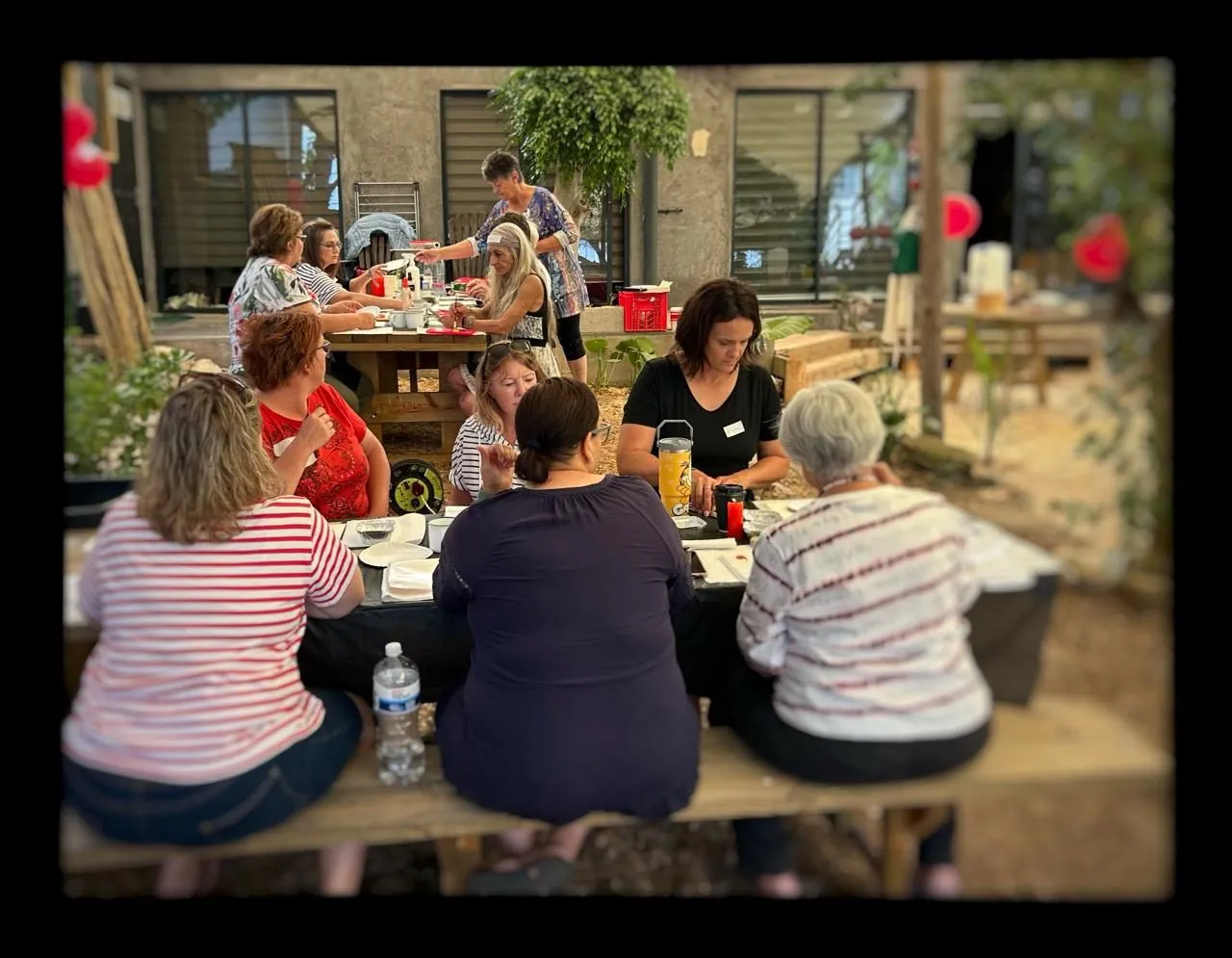 A group of women sitting at tables outdoors under a pergola, seen from behind.