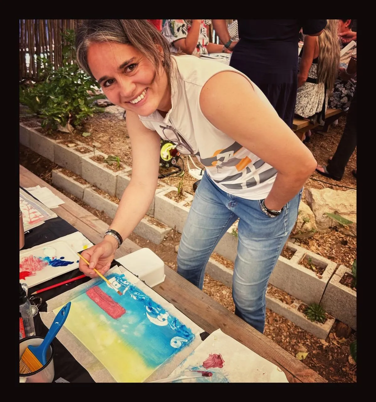 A smiling woman in a sleeveless top paints at a table outdoors, with a block wall behind her.