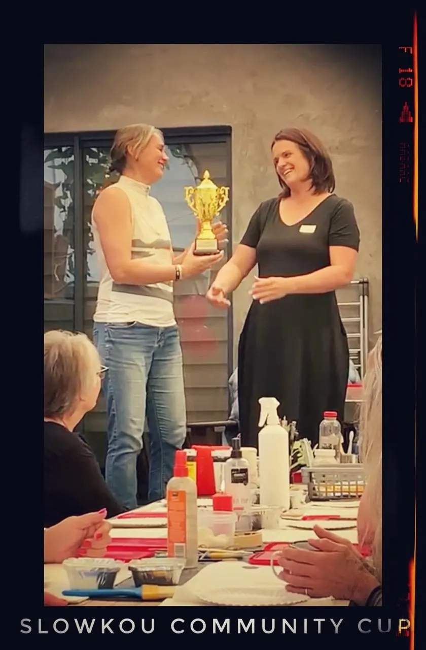 A man in jeans shakes hands with a woman in black who holds a trophy, at a table with bottles, captioned Slowkou community cup.