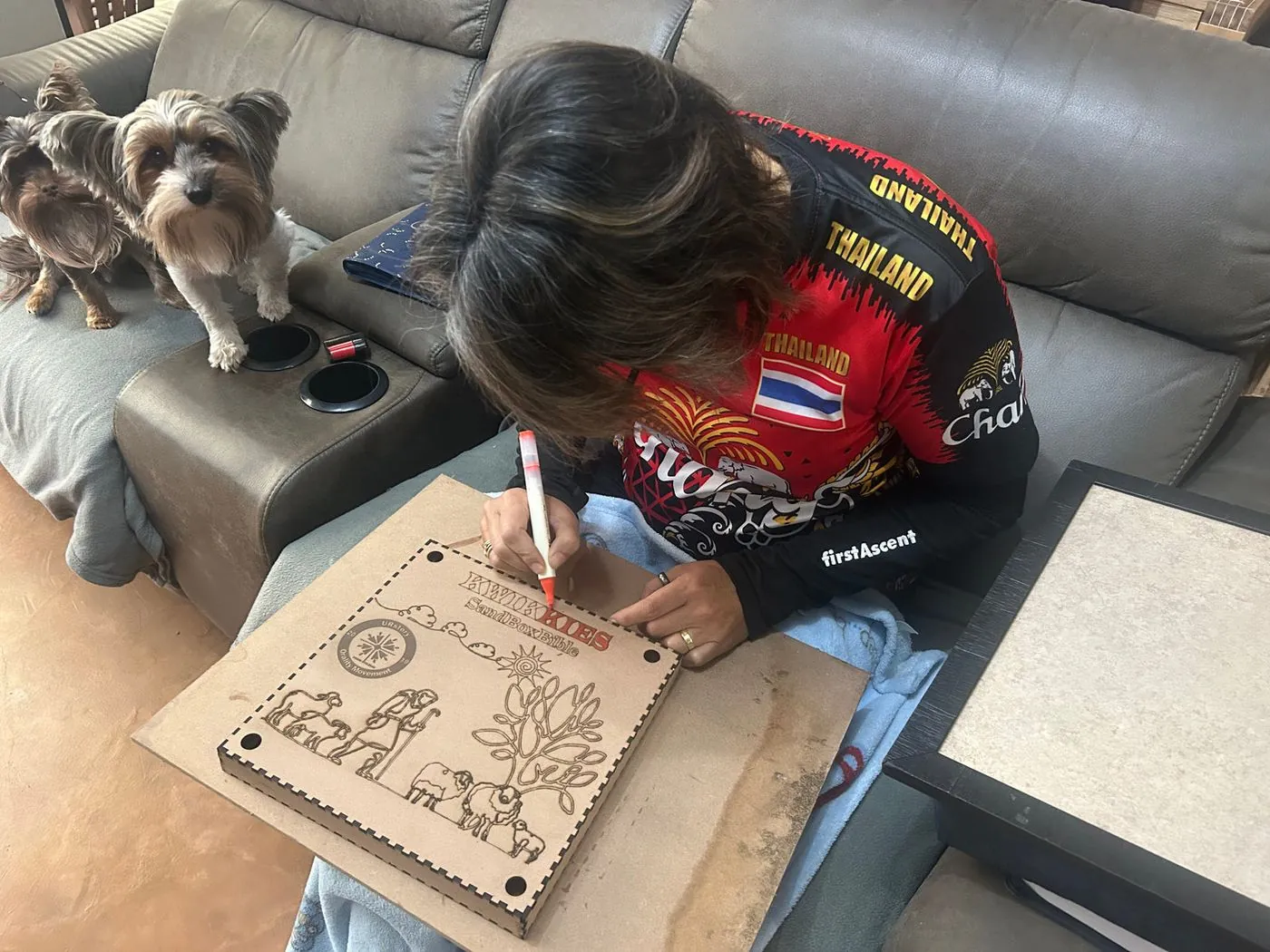 A woman in a riding jacket engraves a wooden board on a sofa, with two small dogs beside her.