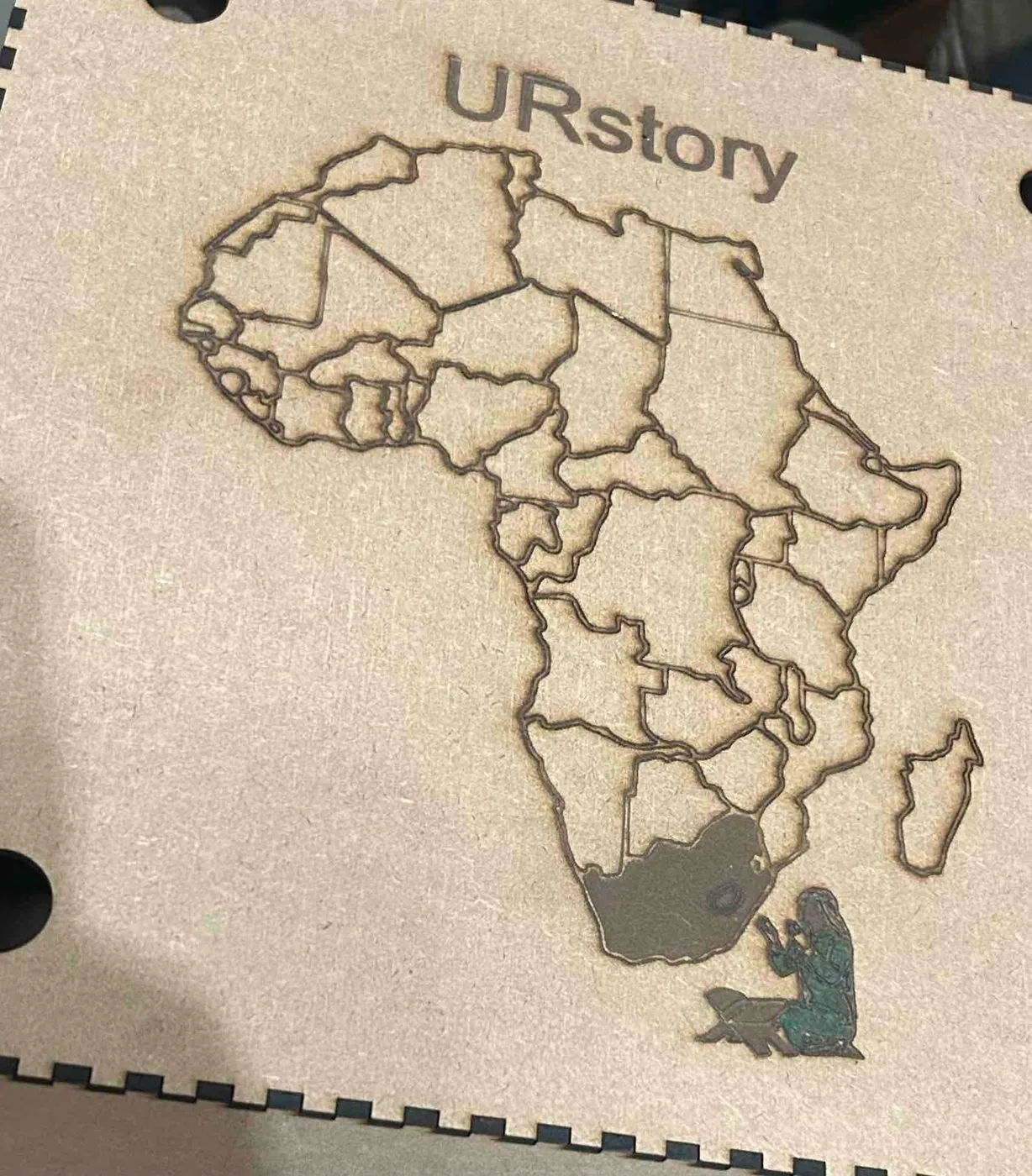 A wooden laser-cut map of Africa with the word URstory engraved on it.