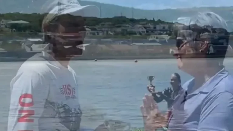 A frame from the post's video: a woman wearing a wetsuit stands in a lagoon holding up a small trophy, with a hillside of houses behind her.