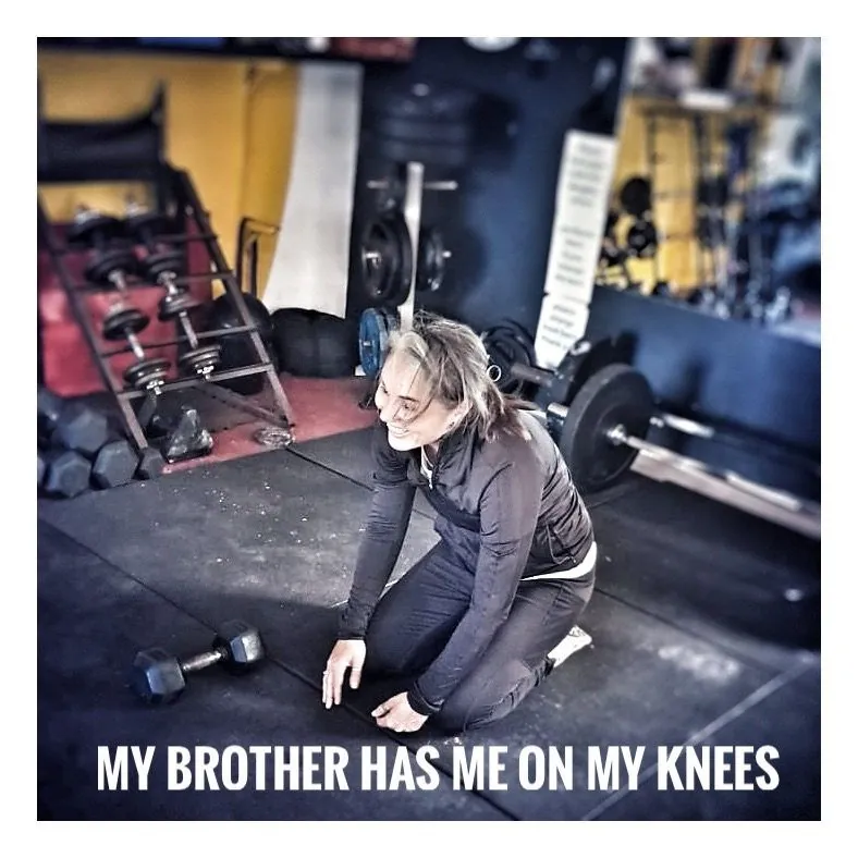 A woman kneeling on a gym floor beside weights, captioned My brother has me on my knees.