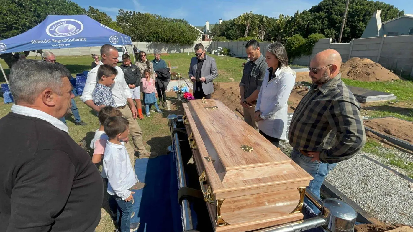 A group of men standing under a blue gazebo beside a wooden coffin at a cemetery.