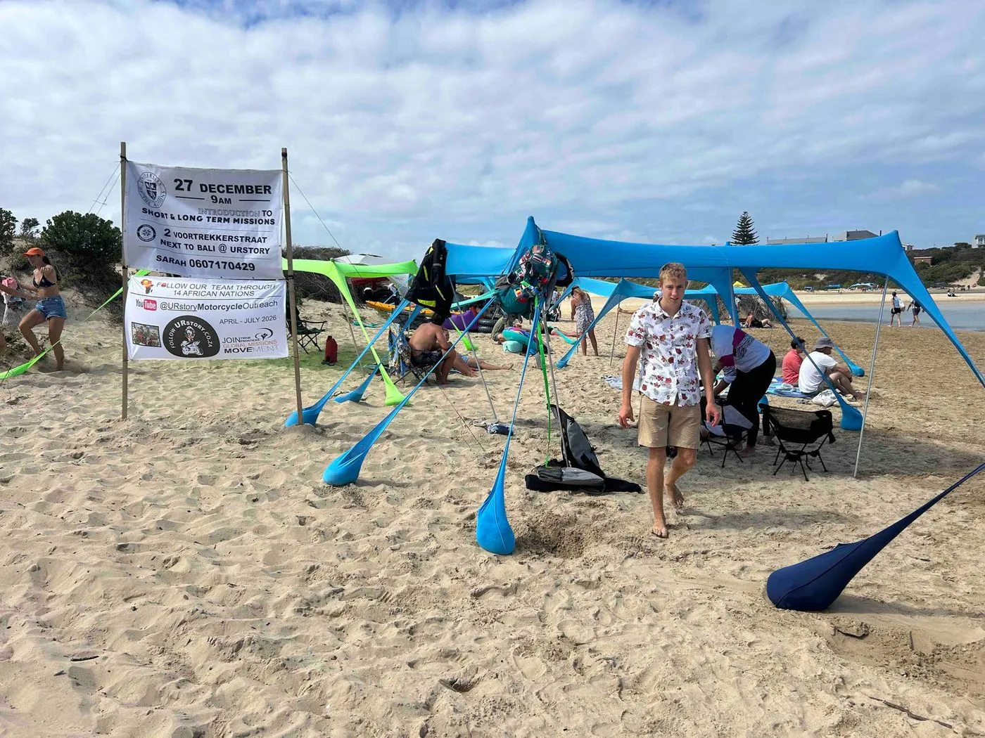 A beach camp with blue and green tarp shelters, a banner on two poles and several people.