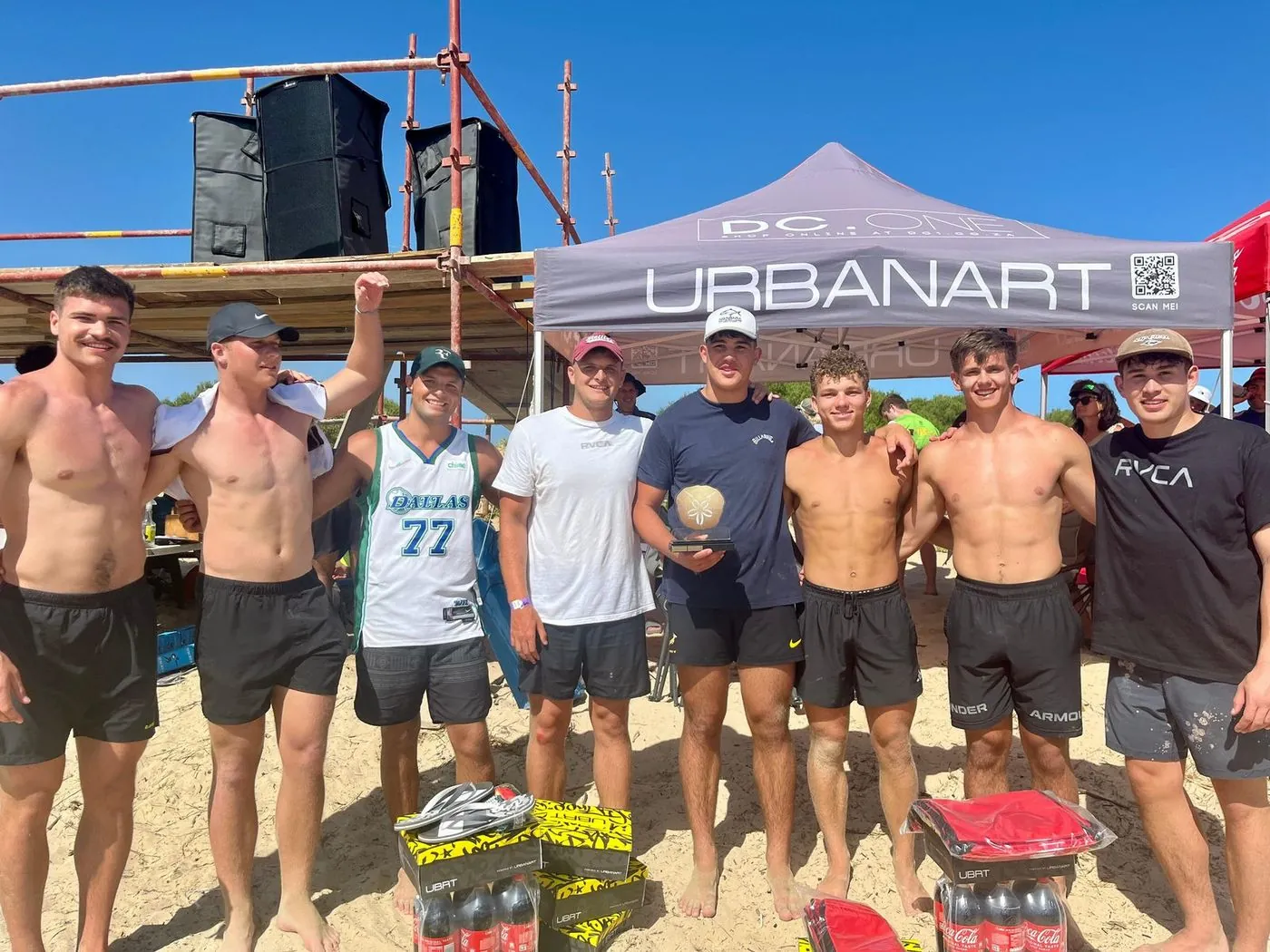 Shirtless men posing in front of a beach marquee marked Urbanart, with other tents behind them.