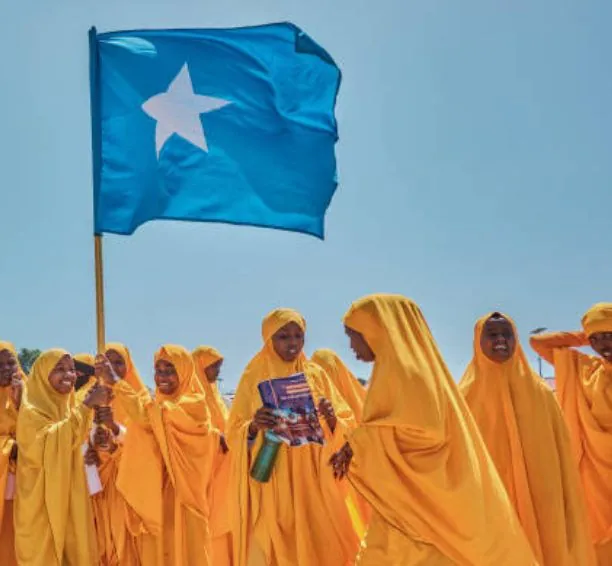 A crowd of women in yellow robes and headscarves, one holding a book, with a pale blue flag with a white star.