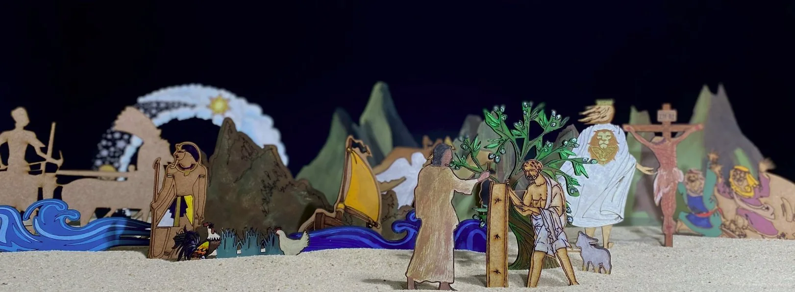 A scene built from flat painted wooden figures standing in pale sand against a black backdrop: blue waves and a boat with a yellow sail, green mountains, an olive tree with two figures beside it, a figure in a white cloak with a lion on its back, and a wooden cross, with a hen and a lamb in the sand.
