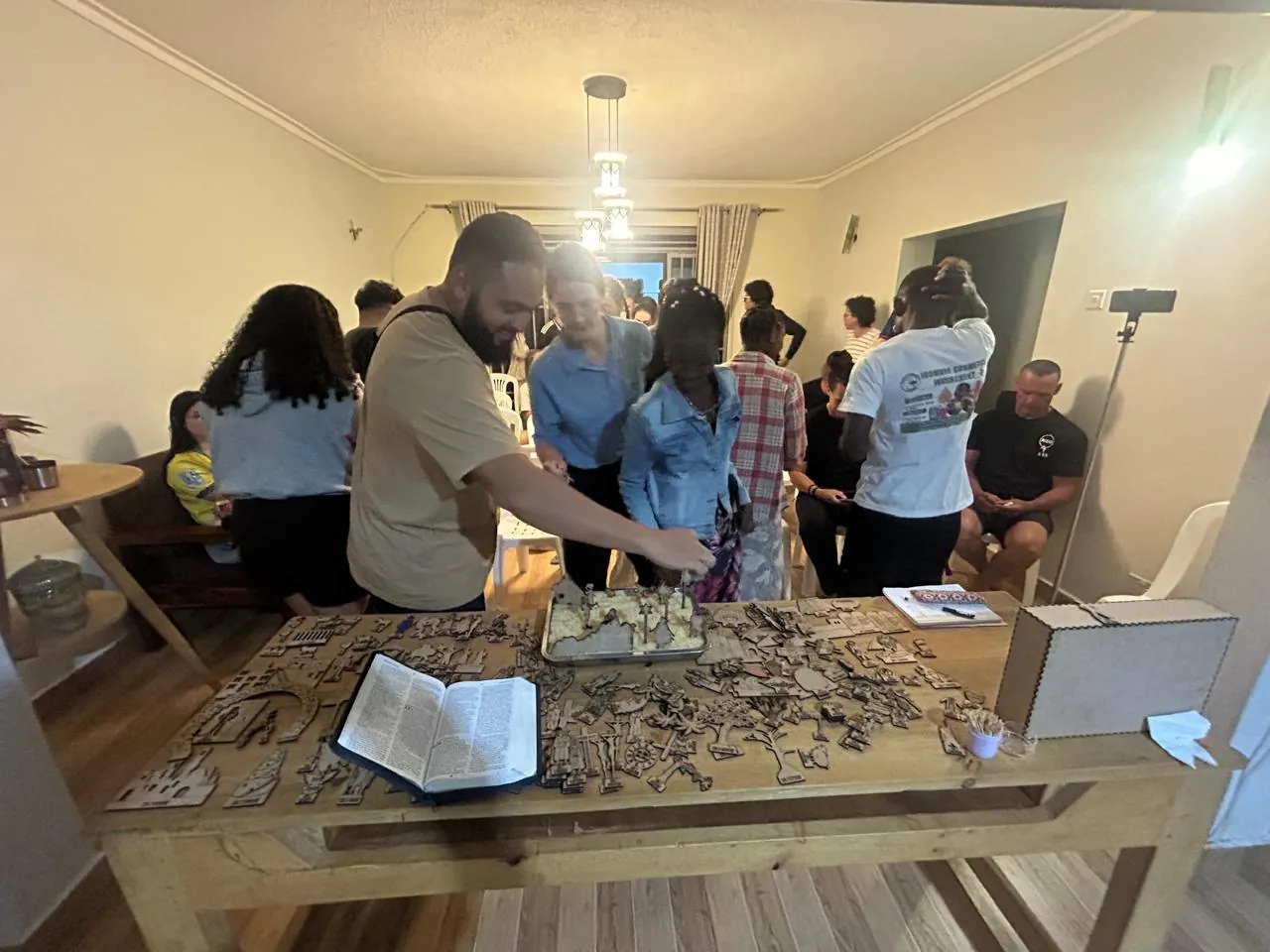A man arranging flat wooden figures on a large table beside an open Bible and a wooden box, while a crowd of people looks on in a bright room.