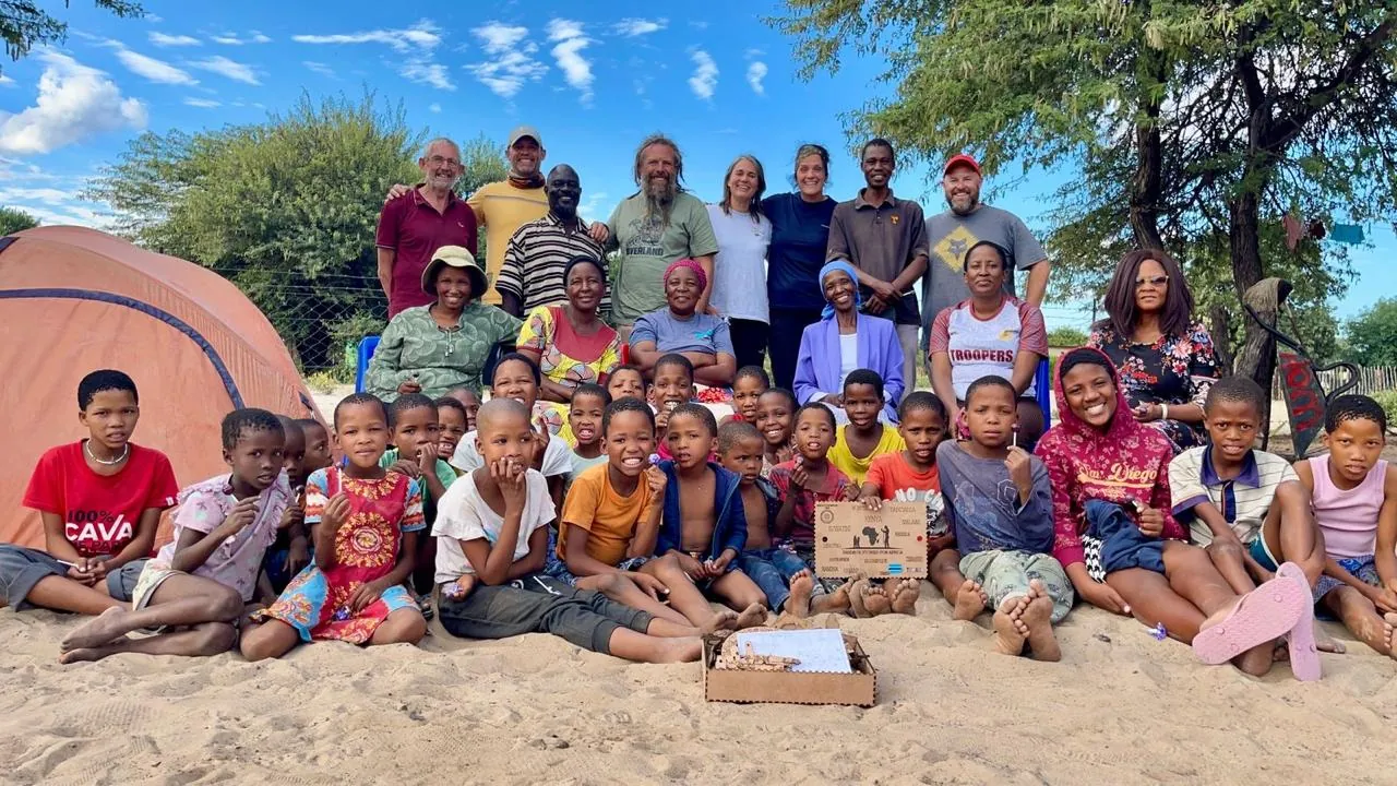 A large group of children sitting on sand in front of two tents, with about twenty adults standing behind them and a small wooden box on the sand in front.