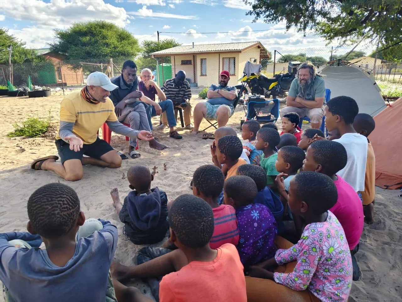 A man in a yellow shirt kneeling on the sand and talking to a circle of children sitting around him, with adults on chairs, a small building and tents behind.