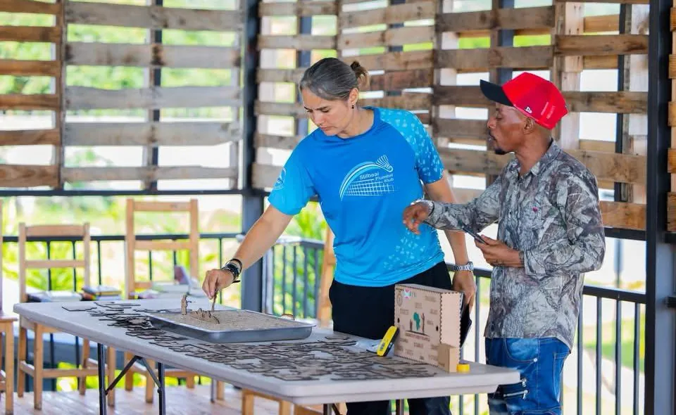 A woman in a blue shirt and a man in a red cap place wooden figures on a long table in an open hall with walls of wooden pallets, next to a small wooden box.
