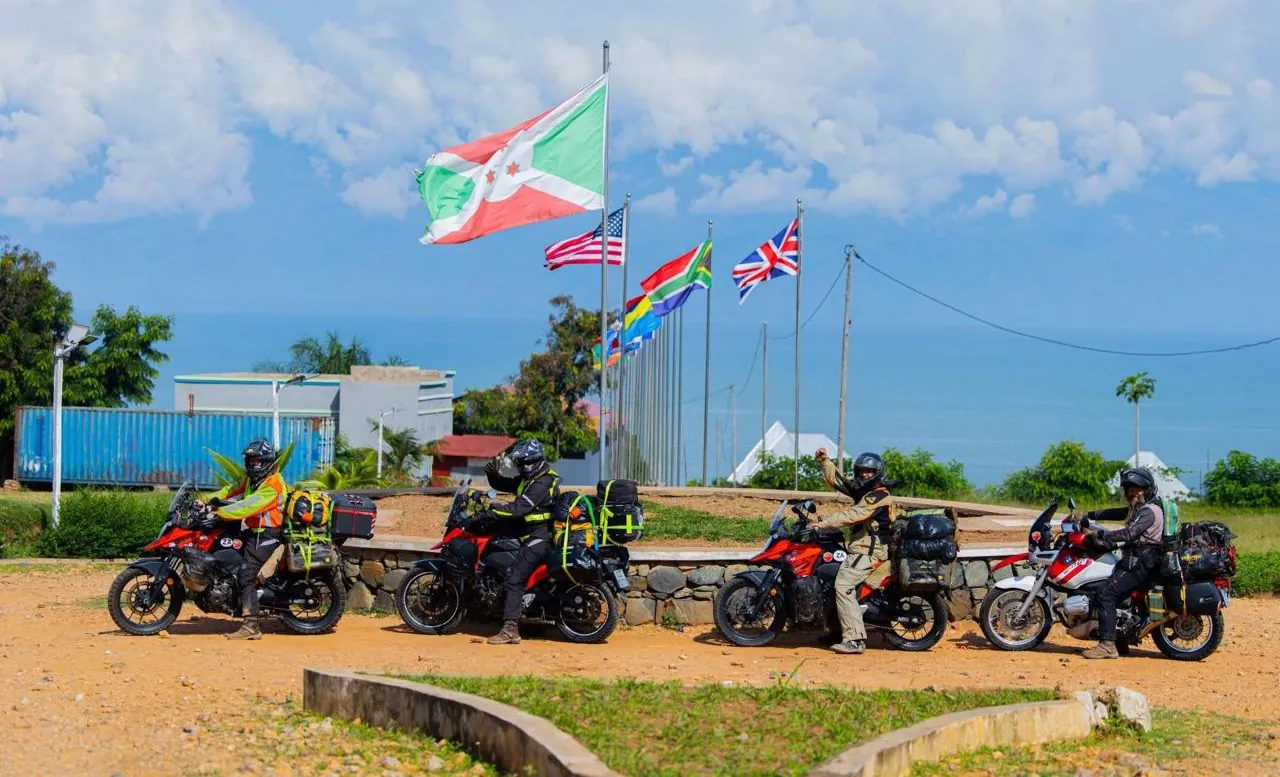 Four motorbikes loaded with luggage parked in a row in front of a line of flags that includes the flag of Burundi, with a lake and a blue sky with clouds behind them.