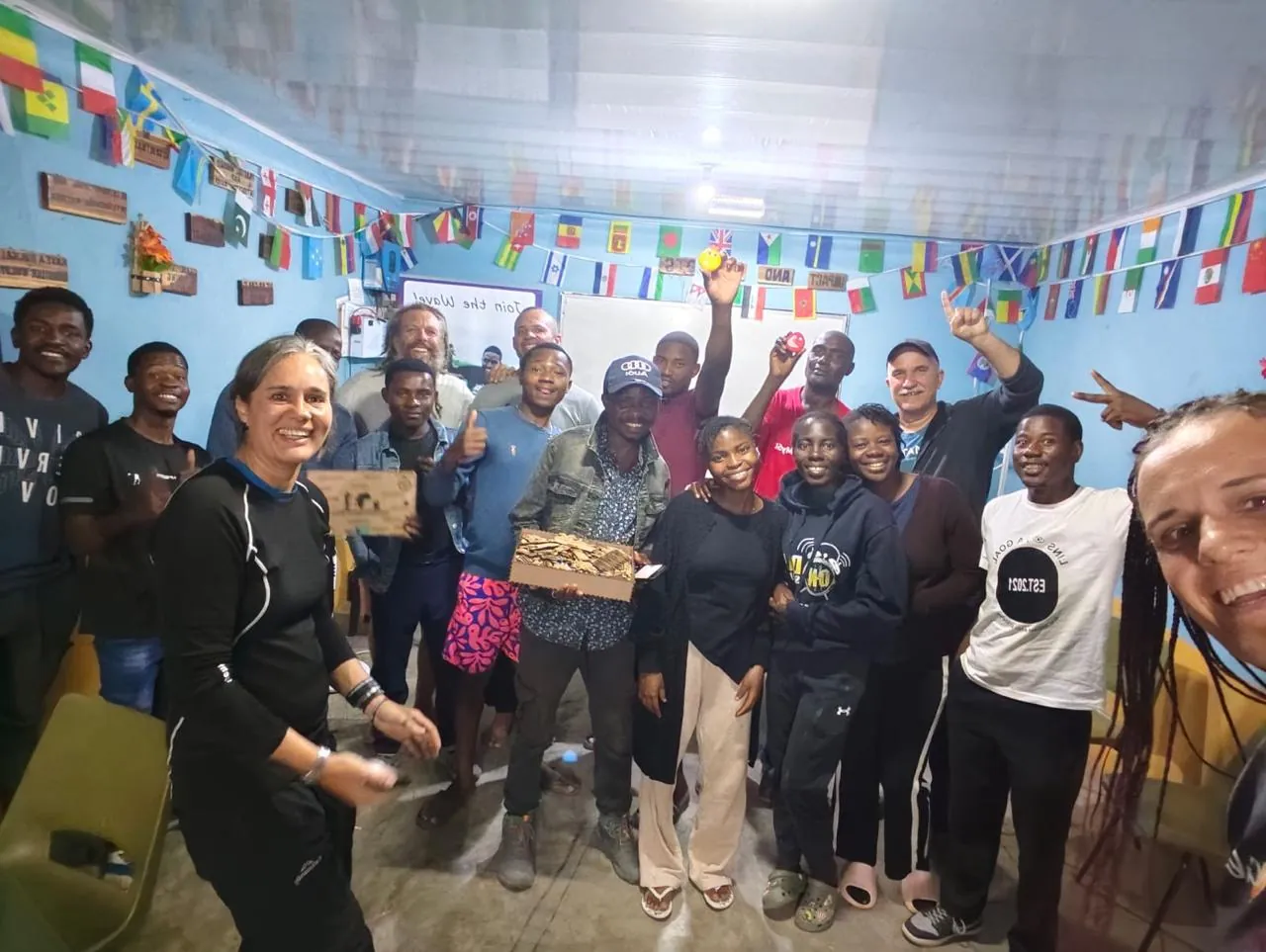 A group of young adults and children crowded into a small blue room hung with national flags, several of them holding up wooden boxes, with a smiling woman in black at the front left.