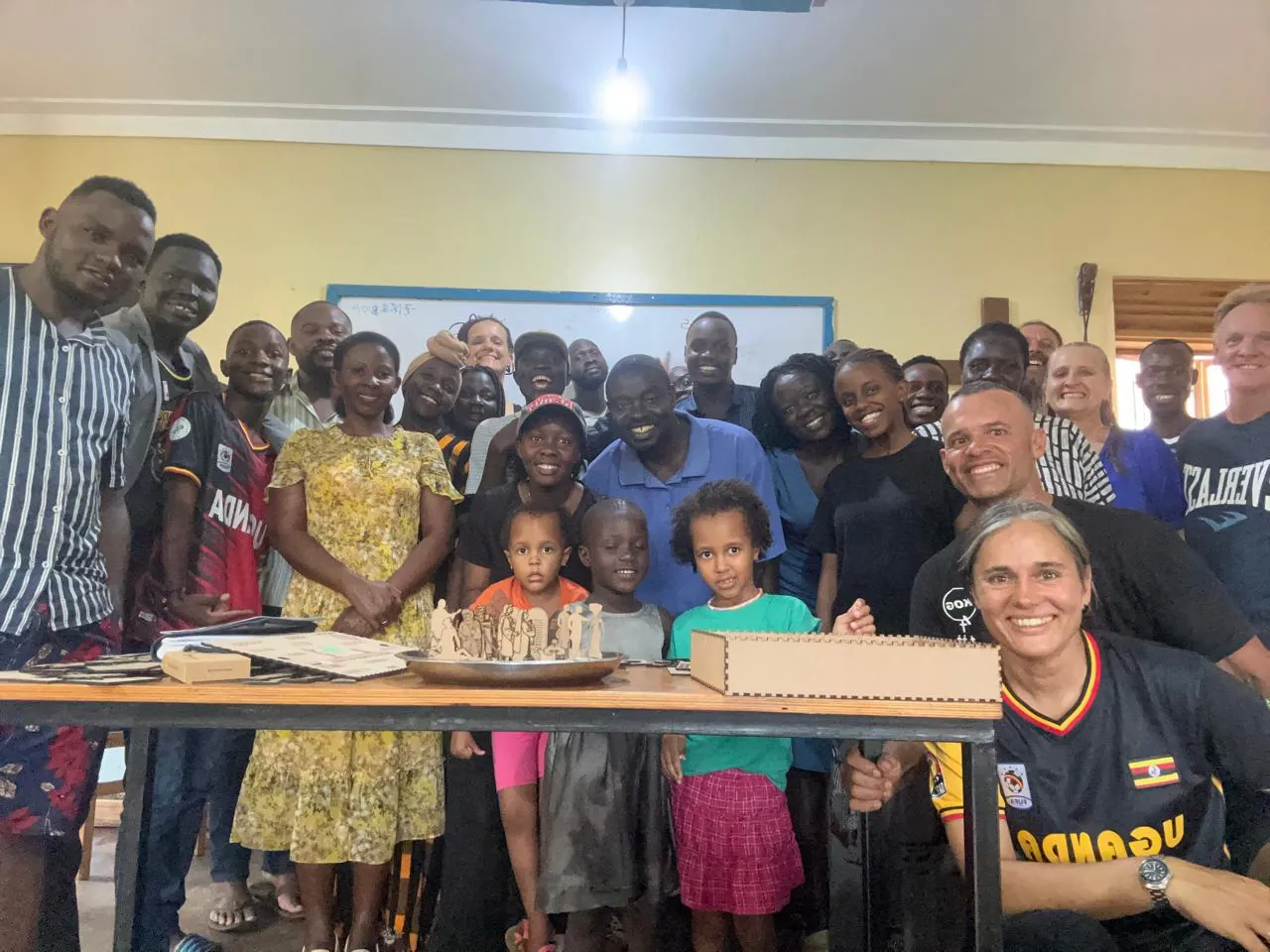 A group of about thirty smiling adults and children crowded together in a classroom, with a woman crouching at the front right beside a table that holds a wooden box and small wooden figures.