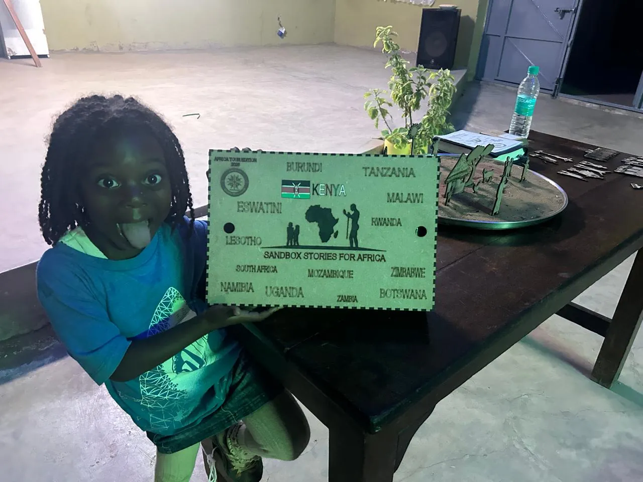A smiling girl poking out her tongue and holding up a green wooden plaque headed Sandbox stories for Africa, with a map of Africa and the names of countries on it, at a dark table.