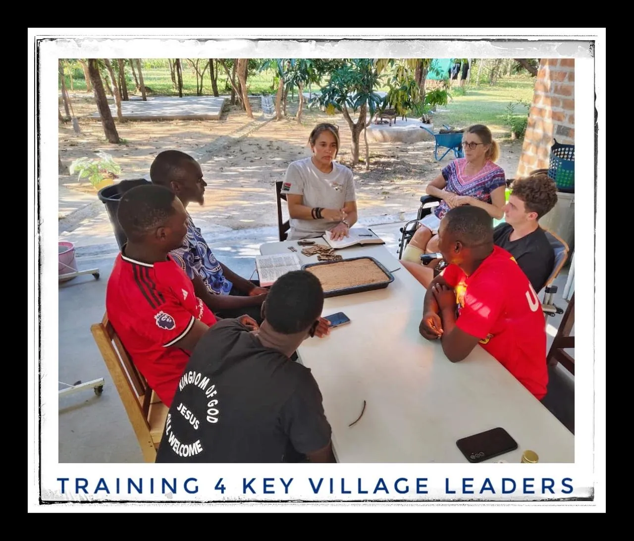 A framed photograph captioned Training 4 key village leaders, showing men and women sitting around a white table in a shaded yard, with small wooden figures on the table.