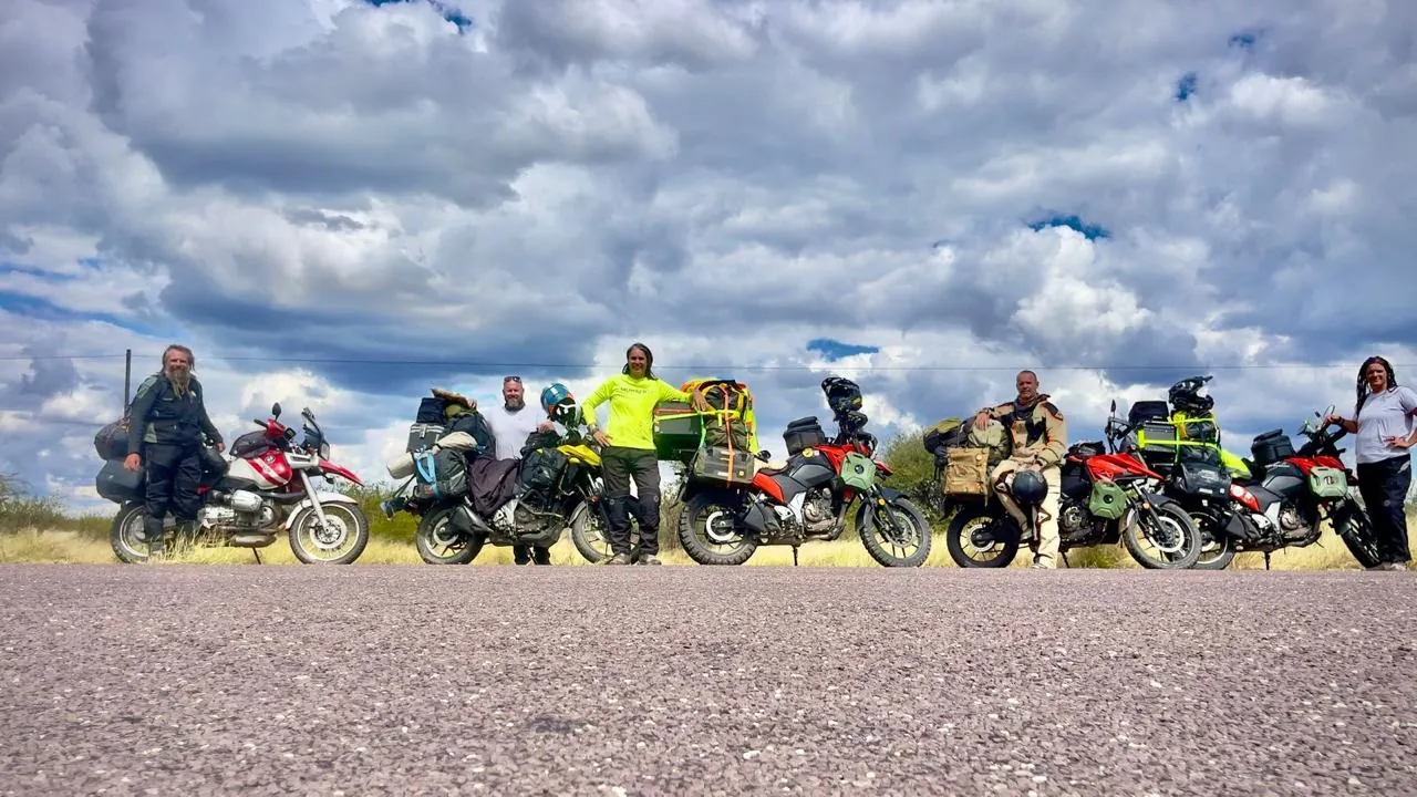 A row of loaded adventure motorbikes and their riders parked on a gravel road under a cloudy blue sky.