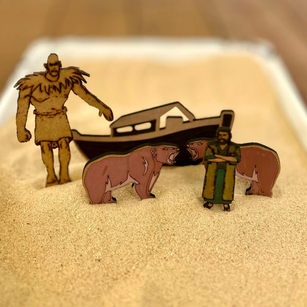 A flat wooden giant, a boat shaped like the ark, two roaring bears and a bearded man in a green robe with folded arms standing in a tray of pale sand.