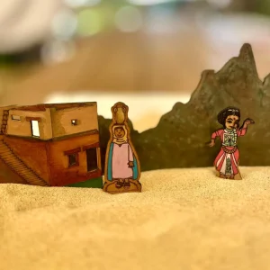 A flat wooden mud-brick house with steps, a woman carrying a water jar on her head and a dancing woman in a red and white costume, with a dark green mountain behind them, standing in sand.