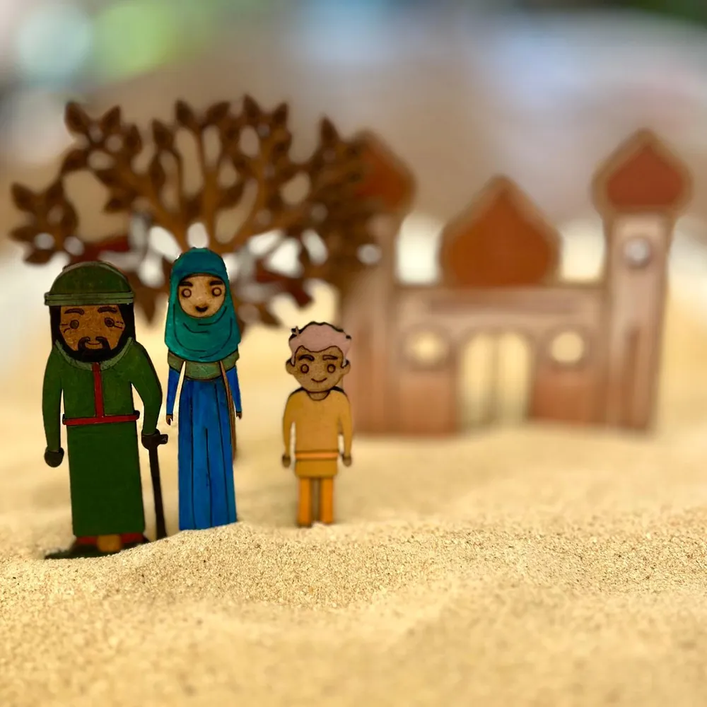 A bearded man in a green robe with a walking stick, a woman in a teal headscarf and a boy, all flat wooden figures, standing in sand with a tree and a mosque behind them.