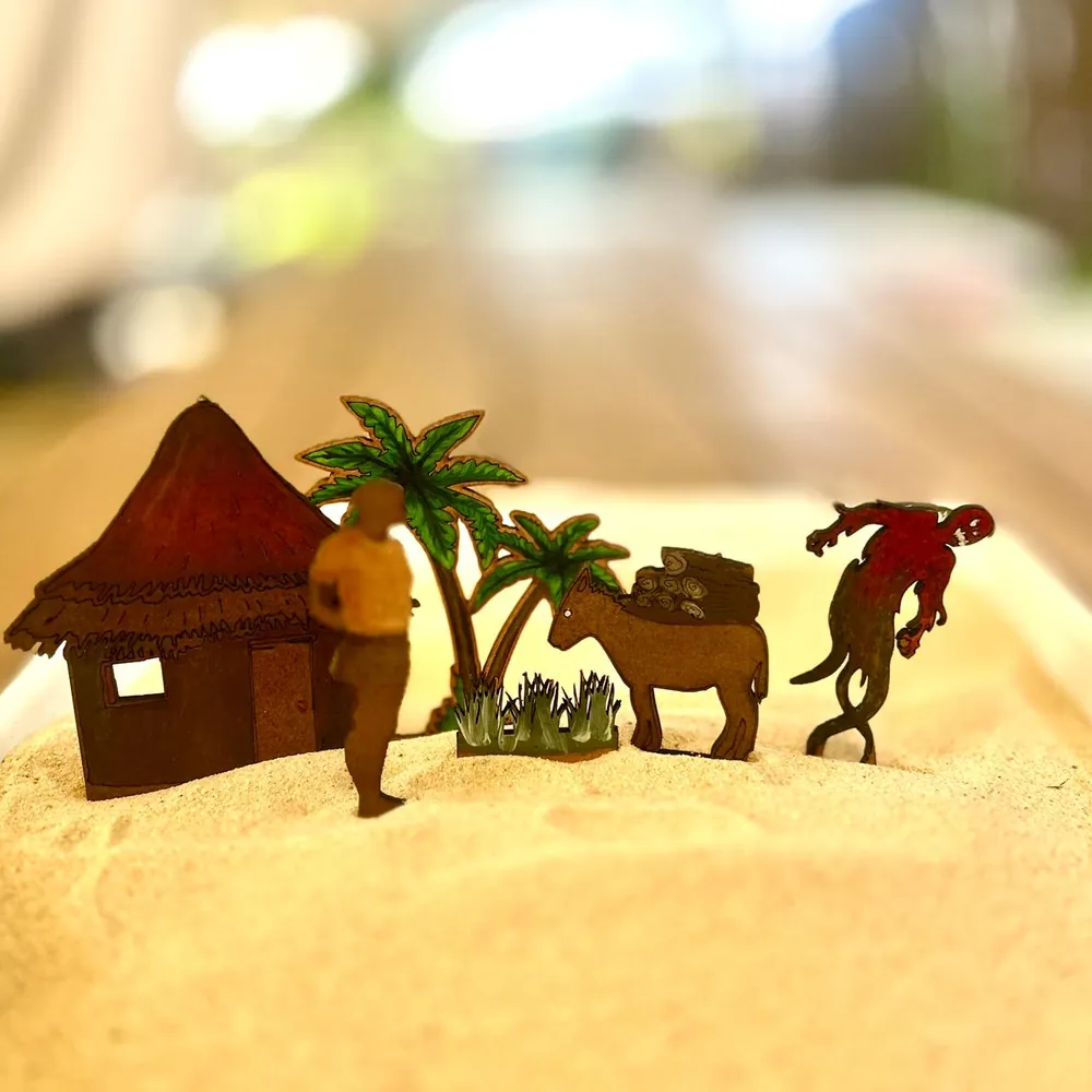A thatched hut, two palm trees, a man in a yellow top, a donkey carrying firewood and a red spirit figure, all flat wooden figures standing in sand.