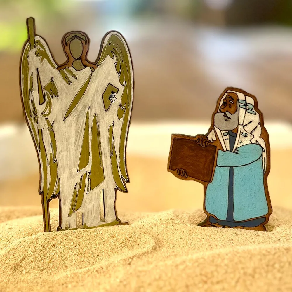 A winged angel with a staff, painted white and olive, and a bearded man in a blue robe and white headcloth holding a dark tablet, both flat wooden figures standing in pale sand.