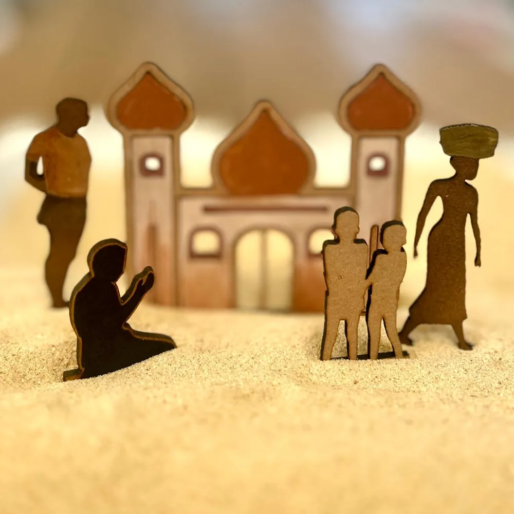 A flat wooden mosque with a kneeling praying figure, a man in an orange shirt, two children and a woman carrying a basket on her head, all standing in sand.