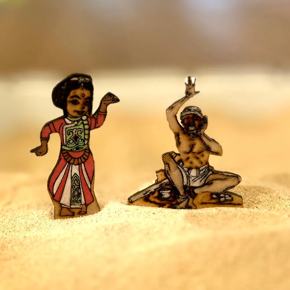 A dancing woman in a red and white costume with a red dot on her forehead and a seated old man with a raised arm and a white head wrap, both flat wooden figures in sand.
