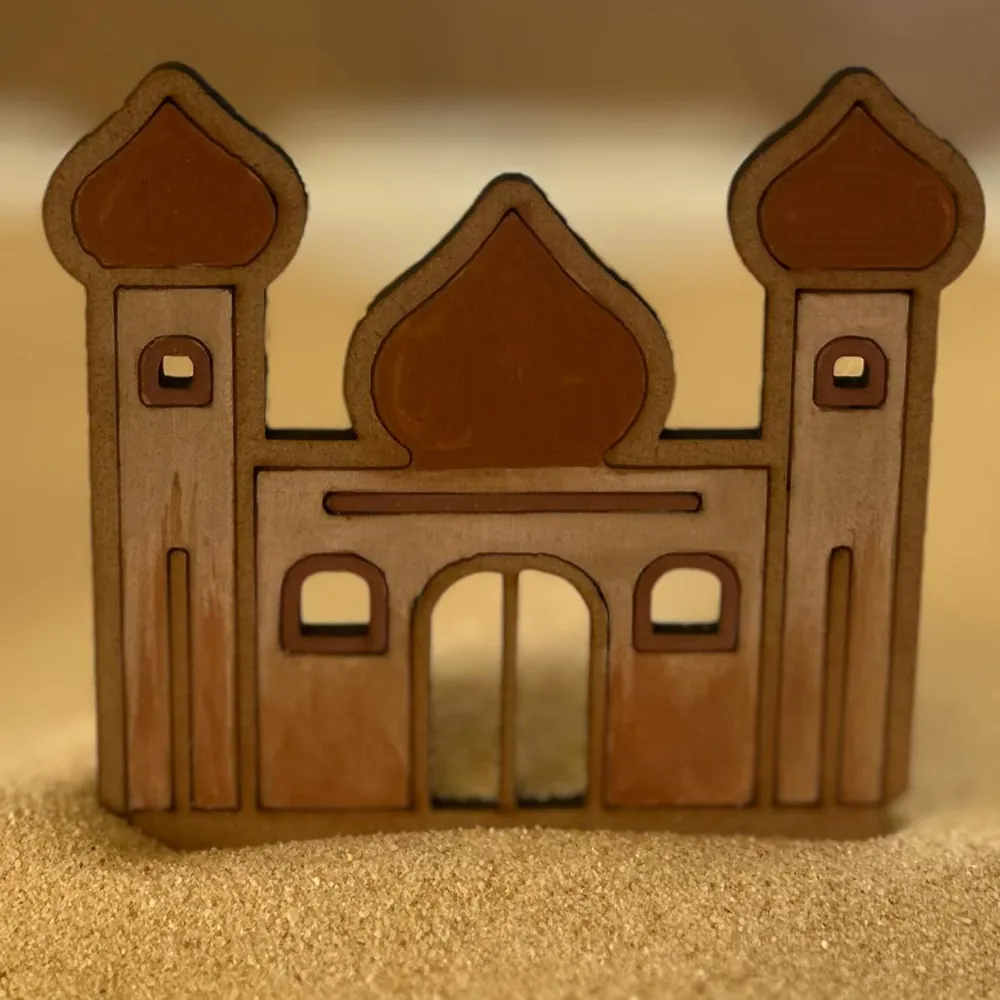 A flat wooden mosque with two onion-domed towers and an arched doorway standing in sand.