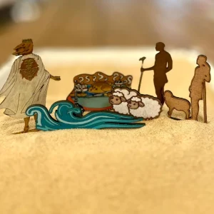 Flat wooden figures in sand: a king in a white cloak seen from behind, a blue wave, two sheep, a table laden with food and two shepherds with staffs.