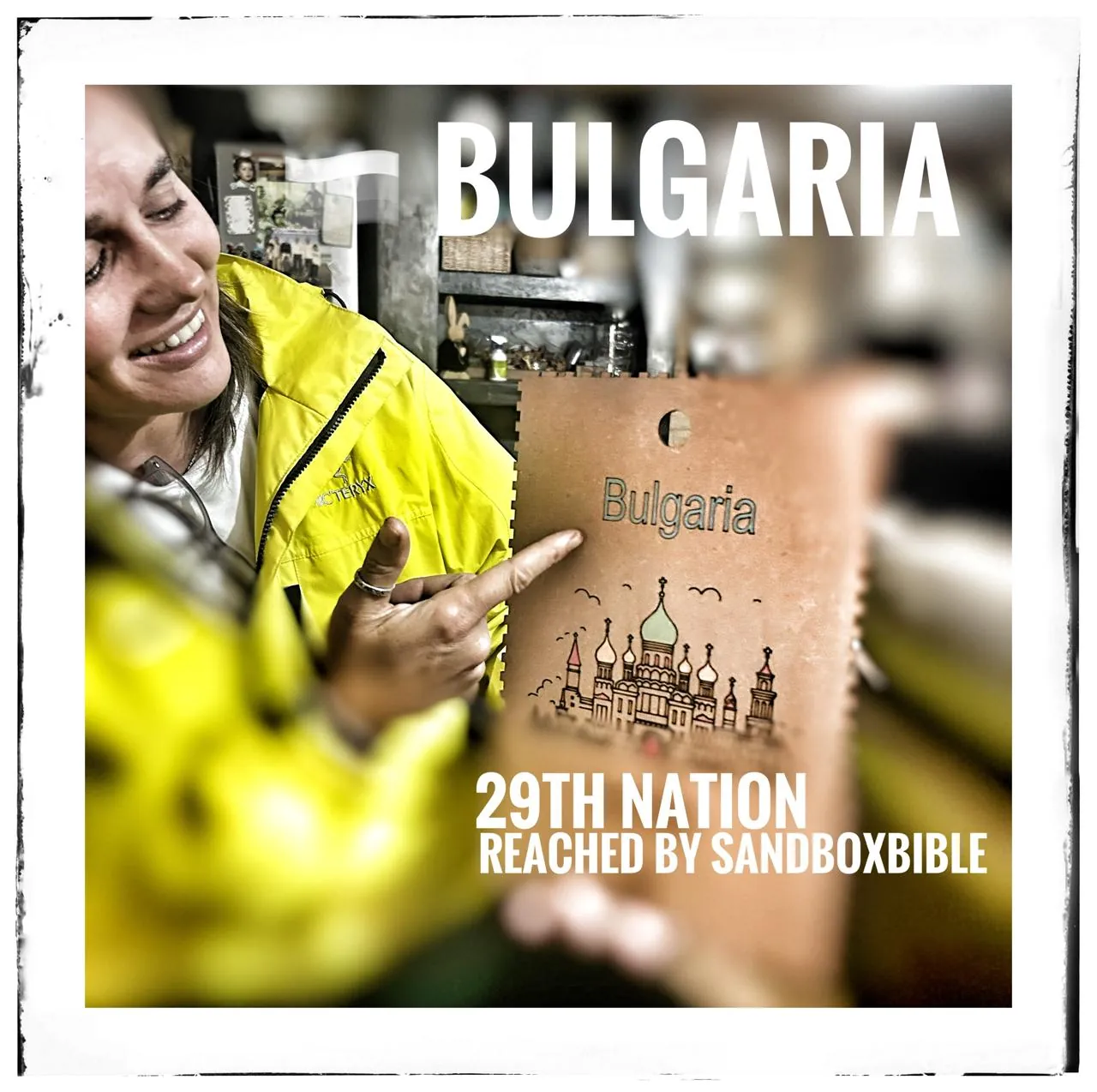A framed print captioned Bulgaria, 29th nation reached by SandBoxBible, showing a smiling woman in a yellow jacket holding up a wooden tag that reads Bulgaria.