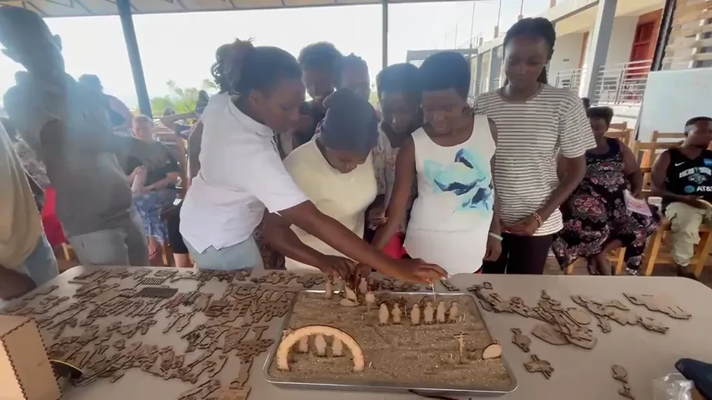 Young women and girls gathered around a table, placing flat wooden figures into a tray of sand, with a crowd watching in an open hall.