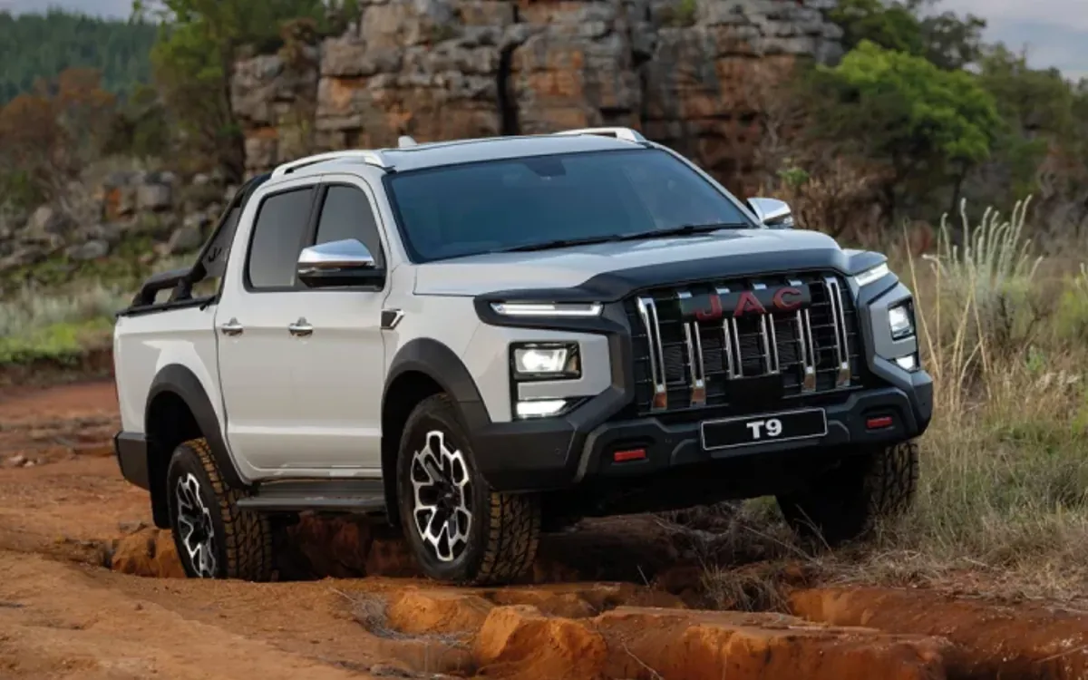 A white JAC double-cab bakkie driving over a rough red dirt track, with rocky hills and dry grass behind it.