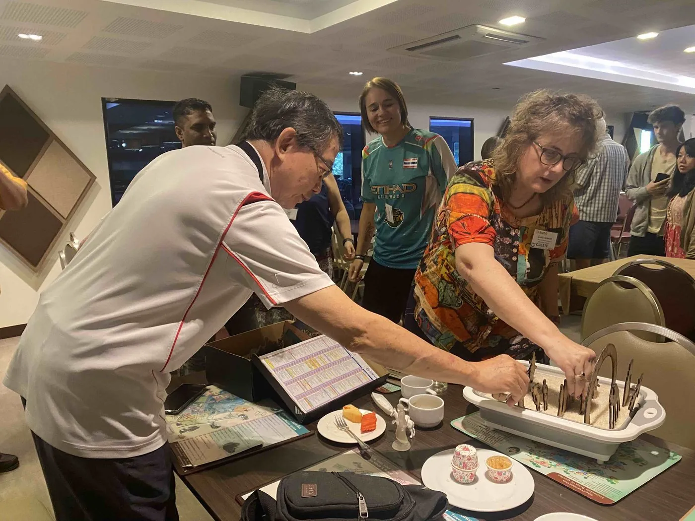 A man in a white shirt and a woman in an orange patterned top reach into a white tray of sand to place small wooden figures, while other people watch behind them in a meeting room.
