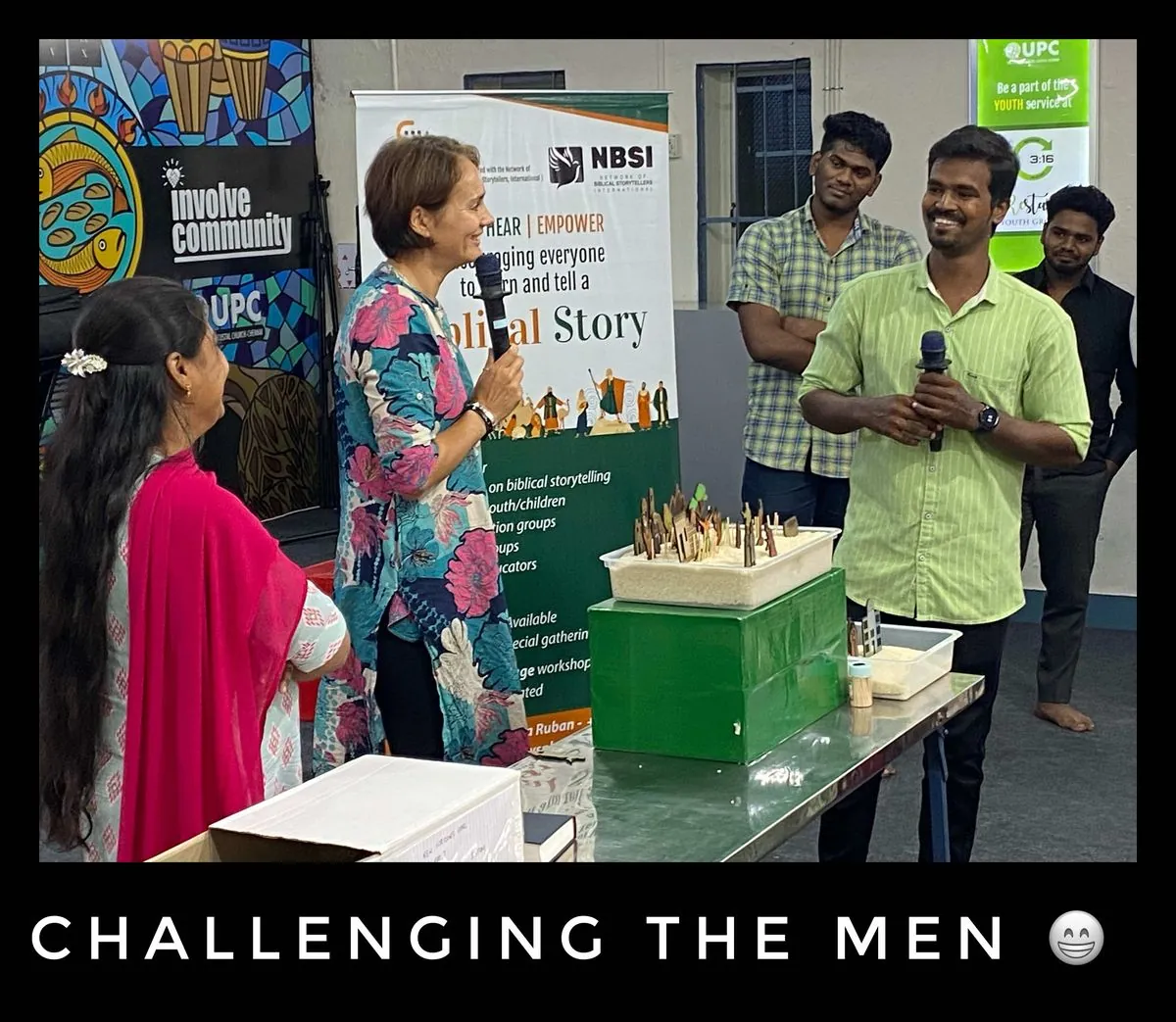 A woman in a patterned jacket speaks into a microphone beside a table with a tray of sand and wooden figures while a smiling man in a green shirt holds a microphone, in front of a banner, with the words Challenging the men below.