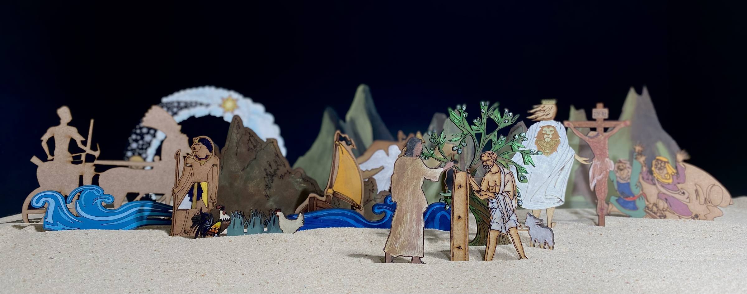 A stage-like scene of flat painted wooden Bible figures standing in pale sand against a dark blue backdrop: a crescent moon and star, mountains, a boat on blue waves, trees, a cross and a figure in white, with small figures in front.