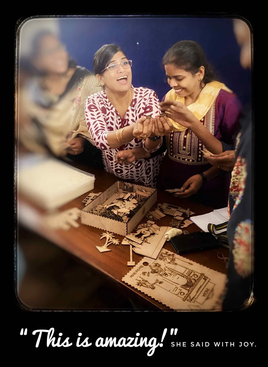 Two women laugh together over a table of wooden figures and open boxes, with the words This is amazing, she said with joy, at the bottom of the picture.