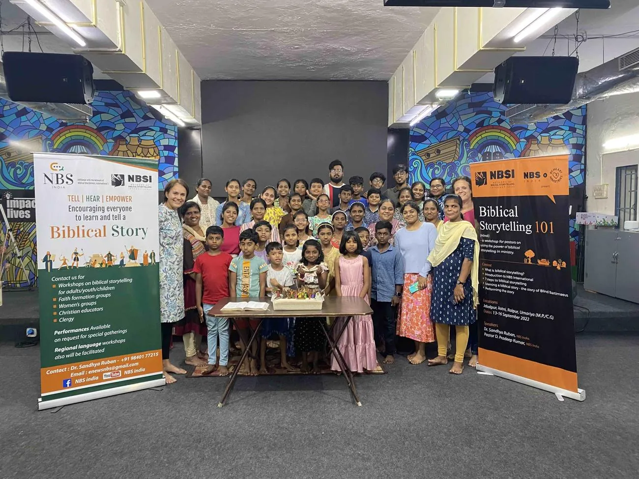 A large group of adults and children stand together in a hall around a table with a tray of sand, between two tall banners that read Tell, Hear, Empower and Biblical Storytelling 101.