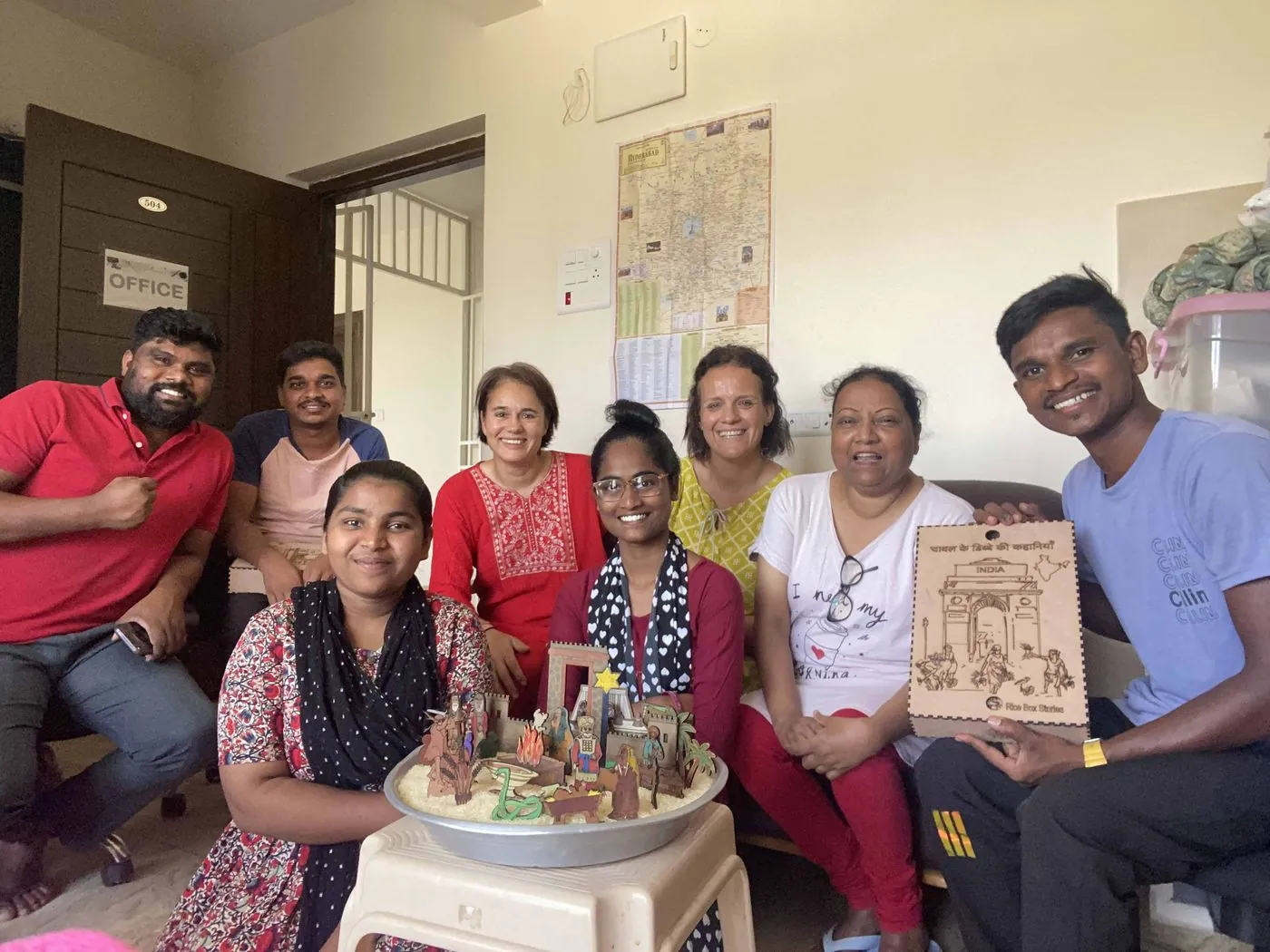 Eight smiling people sit close together in a bright room, holding a tray of sand with painted wooden figures and a carved wooden plaque.