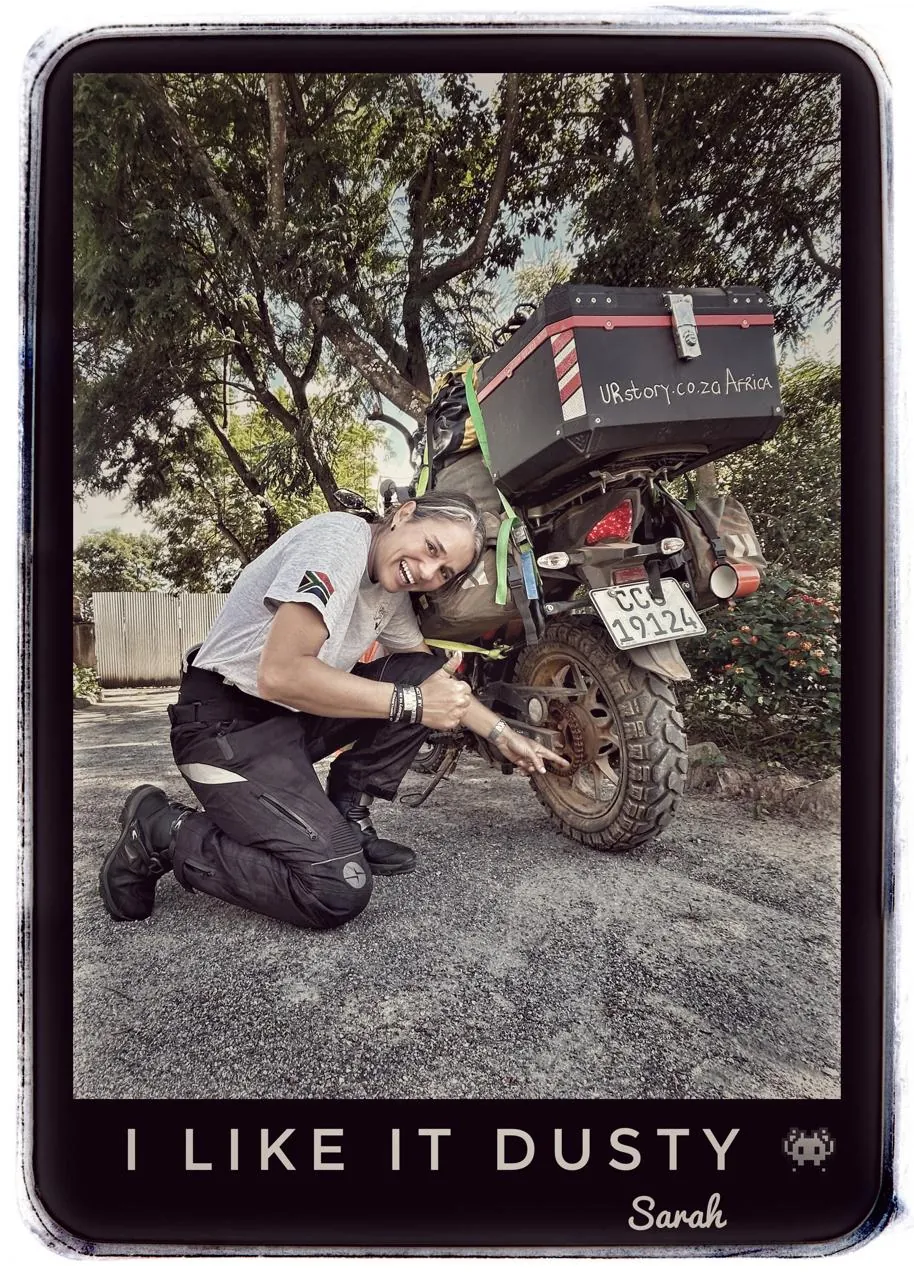 A framed photograph captioned I like it dusty, Sarah, showing a woman in motorcycle gear crouching beside a dusty motorbike with a luggage box, on a road lined with trees.