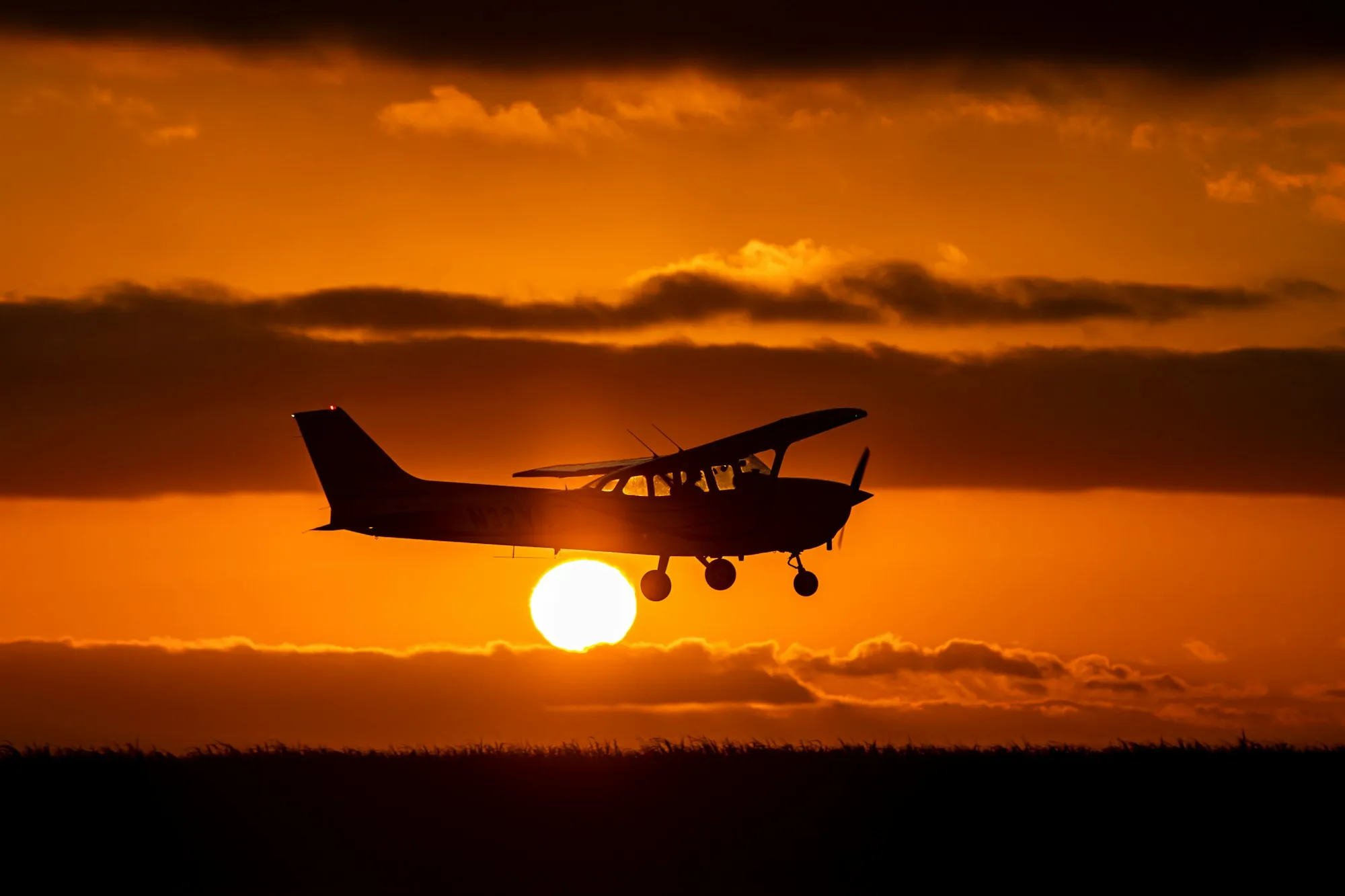 A small single-engine aircraft in silhouette flying low across an orange sunset sky, with the sun just above a dark horizon.