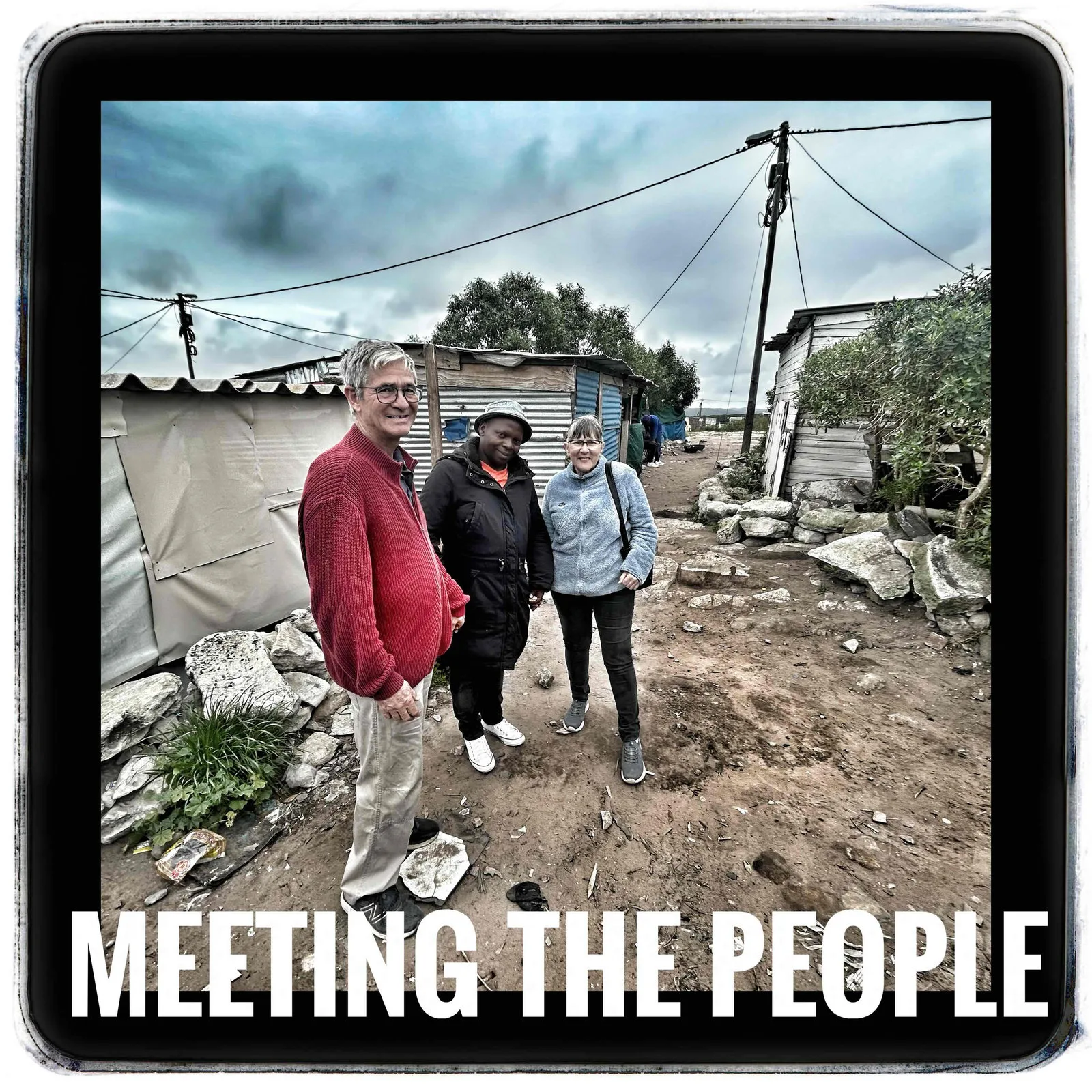 Three people standing on a rocky dirt path between corrugated iron shacks under a cloudy sky, captioned Meeting the people.