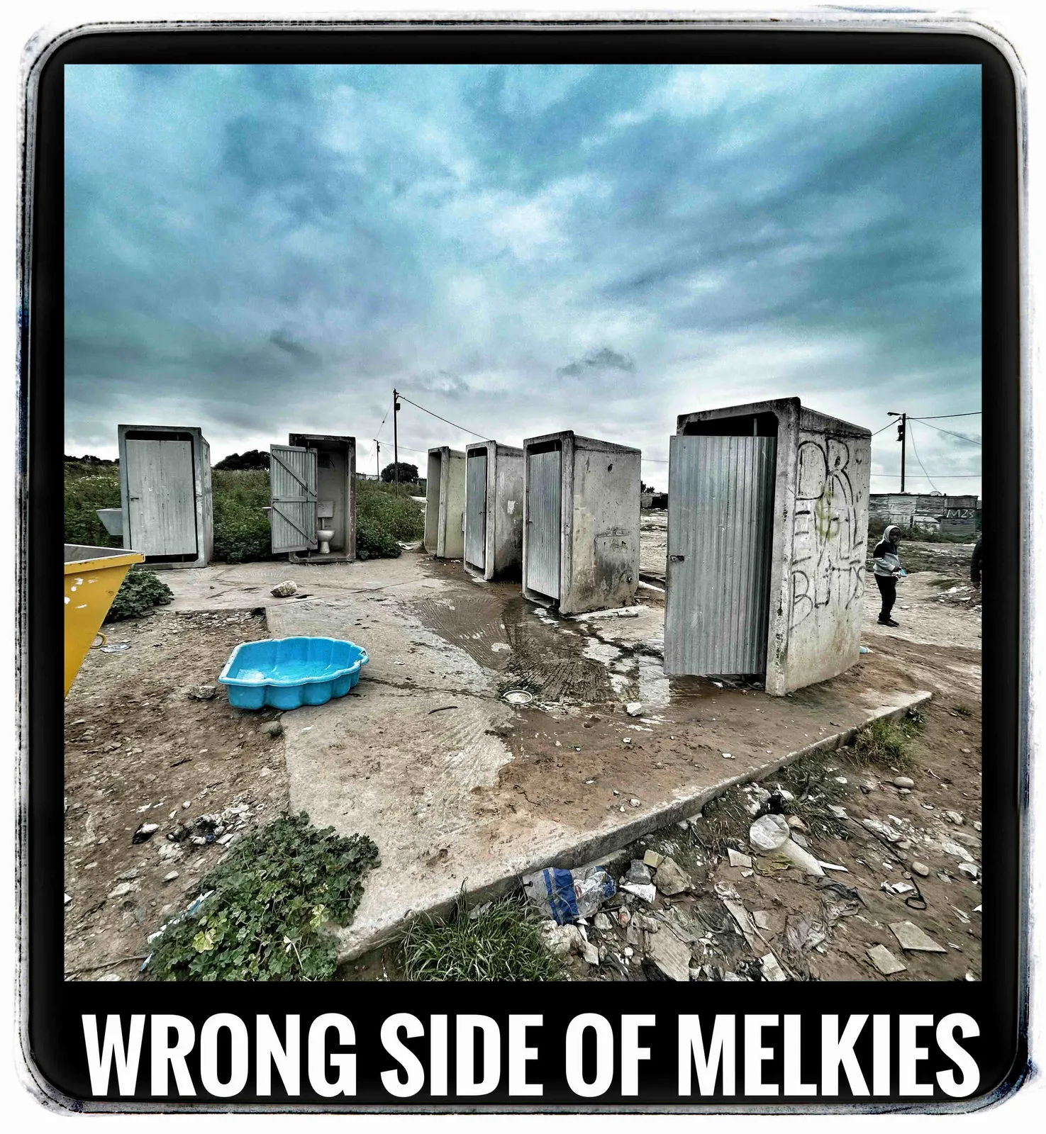 A row of metal-clad toilets on rocky ground beside a blue paddling pool under a cloudy sky, captioned Wrong side of Melkies.